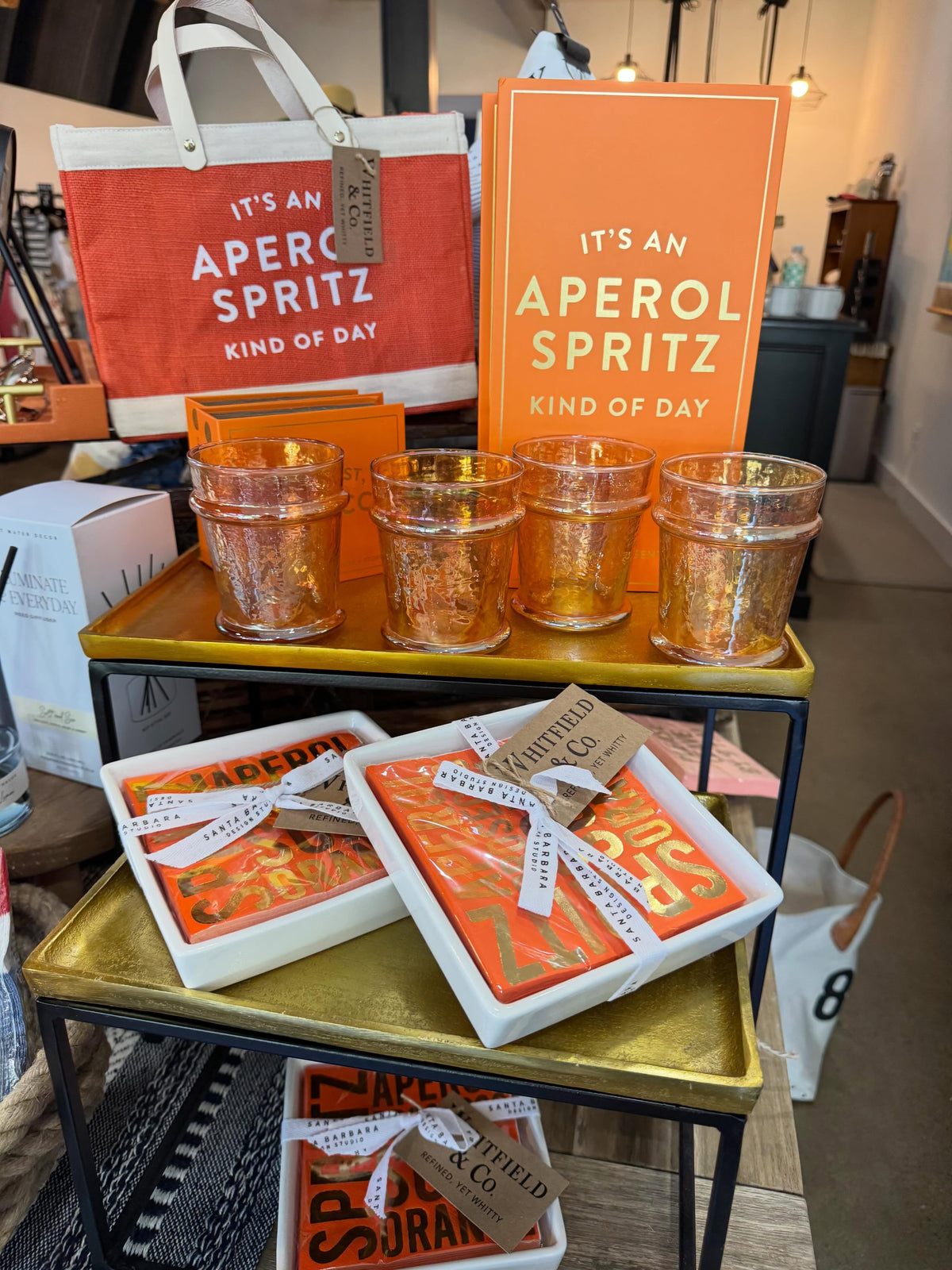 A retail display featuring Aperol Spritz themed merchandise, including orange and red tote bags, matching greeting cards, and four copper-toned glittery cocktail glasses on a gold-trimmed black metal shelf. Several boxed Aperol Spritz mix packets are also arranged on the shelves.