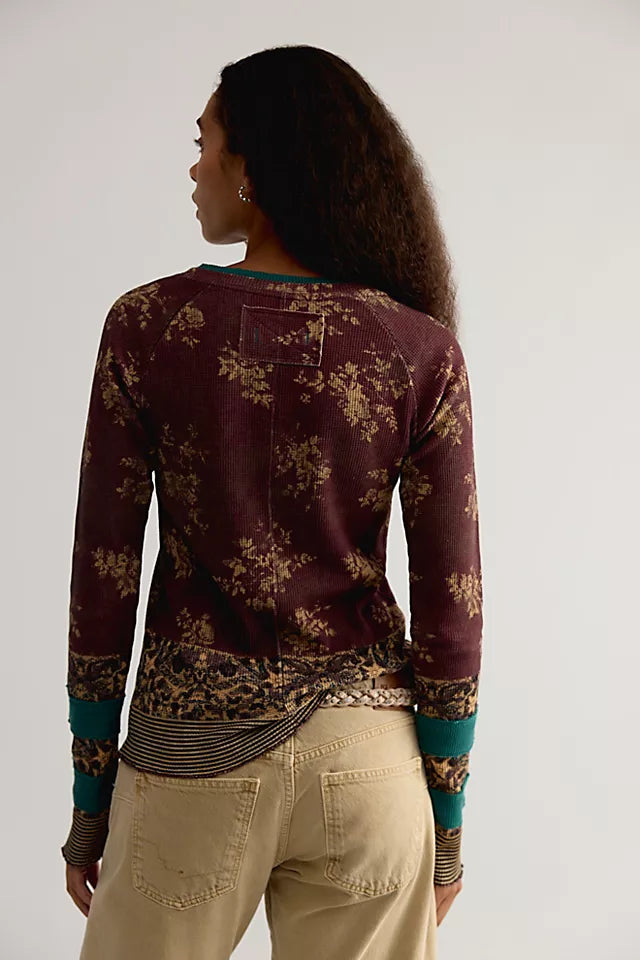 A woman shown from the back wearing a long-sleeve maroon top with a beige floral pattern, teal and black striped cuffs, and beige pants against a plain light gray background.