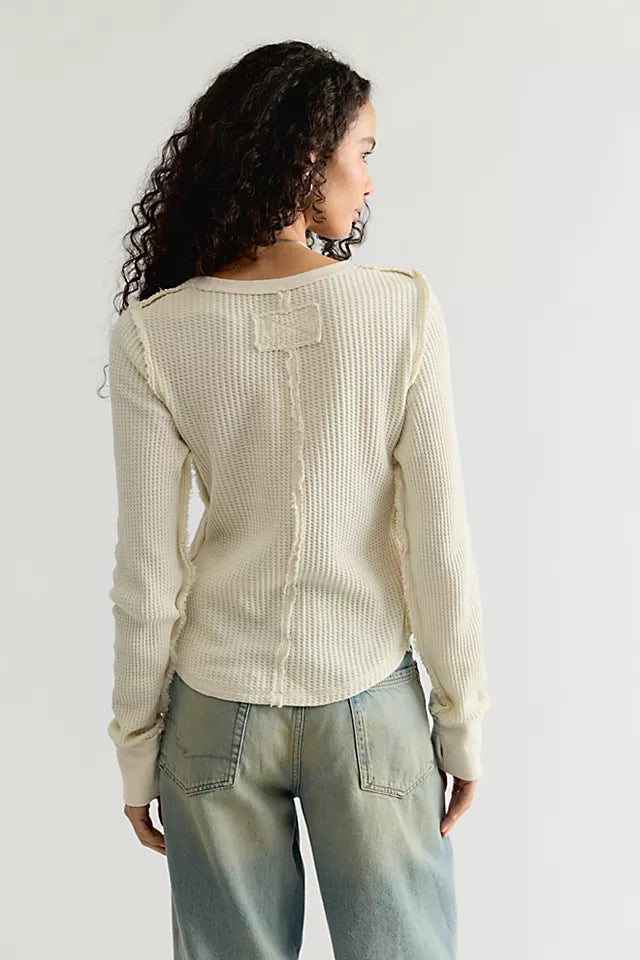 A woman shown from the back wearing a cream ribbed knit sweater with shoulder details and light-wash jeans, against a plain light gray studio background.