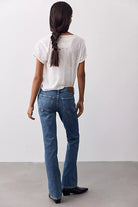 A woman viewed from behind wearing blue straight-leg jeans, a white short-sleeve top, and black boots against a plain light gray studio background.