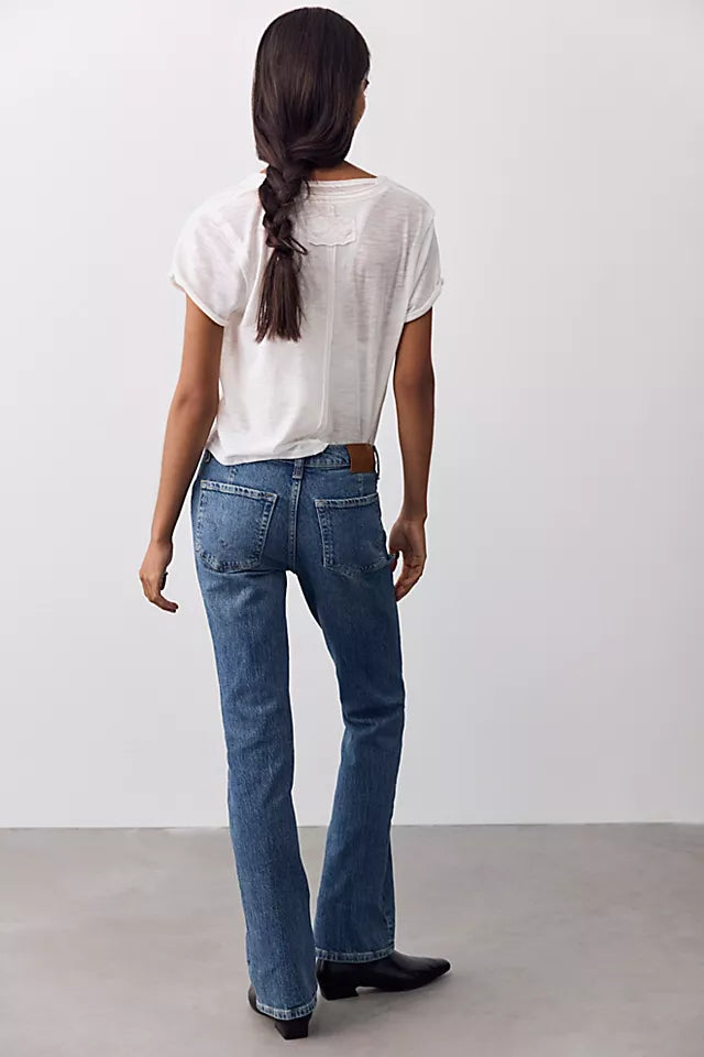 A woman viewed from behind wearing blue straight-leg jeans, a white short-sleeve top, and black boots against a plain light gray studio background.