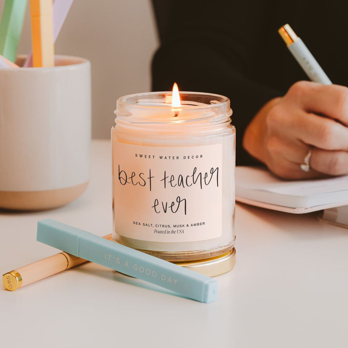 Candle labeled 'best teacher ever' on a desk with writing utensils and a person writing.