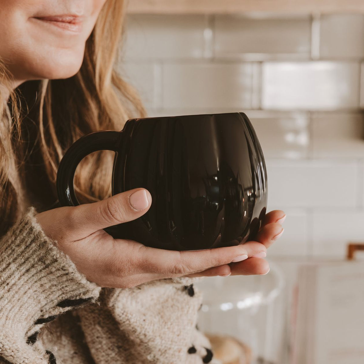A person in a beige knit sweater holds a glossy black ceramic mug in a warm, softly lit kitchen setting with white subway tiles in the background.
