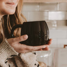 A person in a beige knit sweater holds a glossy black ceramic mug in a warm, softly lit kitchen setting with white subway tiles in the background.