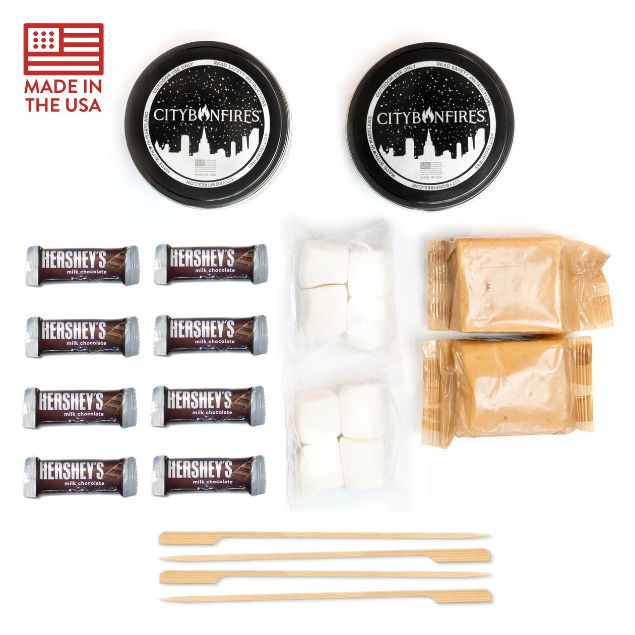City of Fires marshmallow s'mores kit components laid out on a white background, including two black-labeled tins, Hershey's chocolate bars, marshmallows, peanut butter packets, wooden skewers, and a 'Made in the USA' flag graphic.