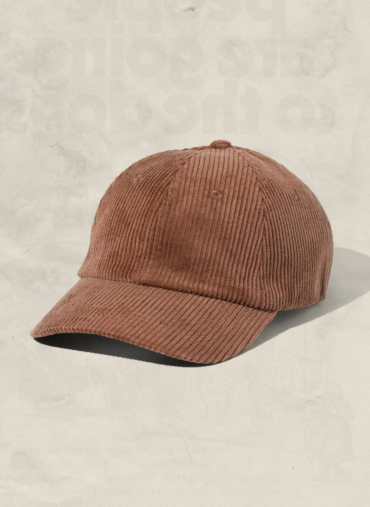 A brown corduroy baseball cap with a curved brim, photographed on a textured light beige background.