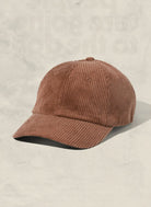 A brown corduroy baseball cap with a curved brim, photographed on a textured light beige background.