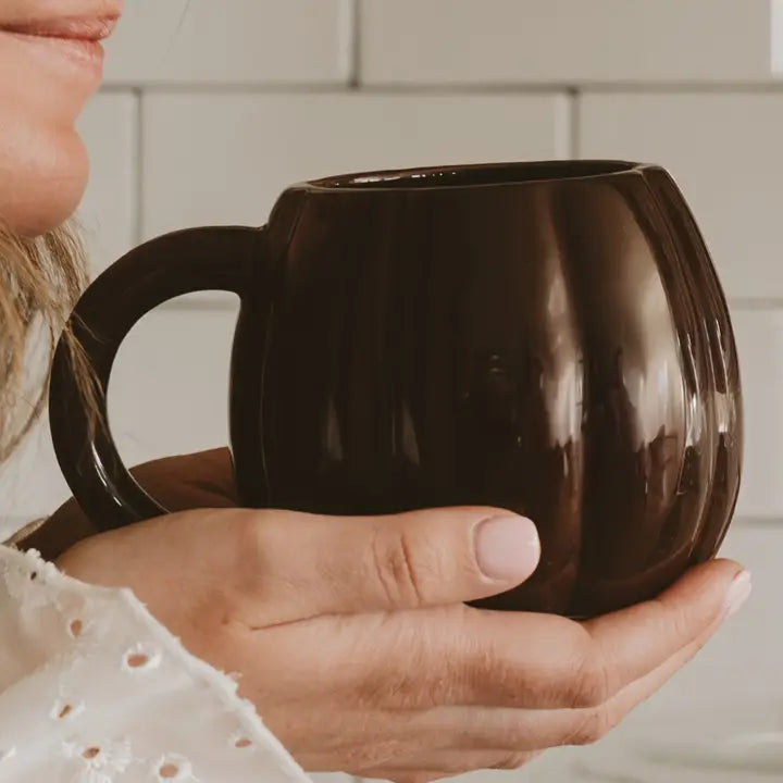 A person holding a glossy dark brown ceramic mug with a rounded, ribbed shape and a curved handle, set against a soft-focus white tiled background.
