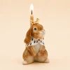 A novelty candle shaped like a small brown bunny wearing a white bow tie, with a lit flame on top, set against a plain beige background.