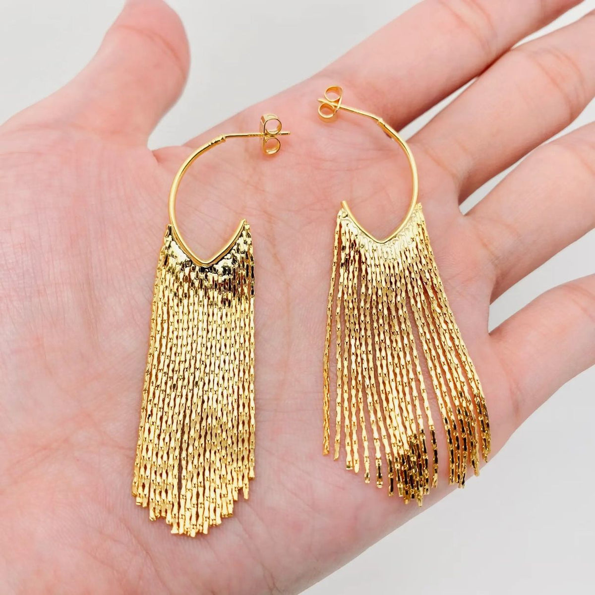 A pair of gold hoop earrings with long, textured fringe, displayed on an open hand against a light gray background.