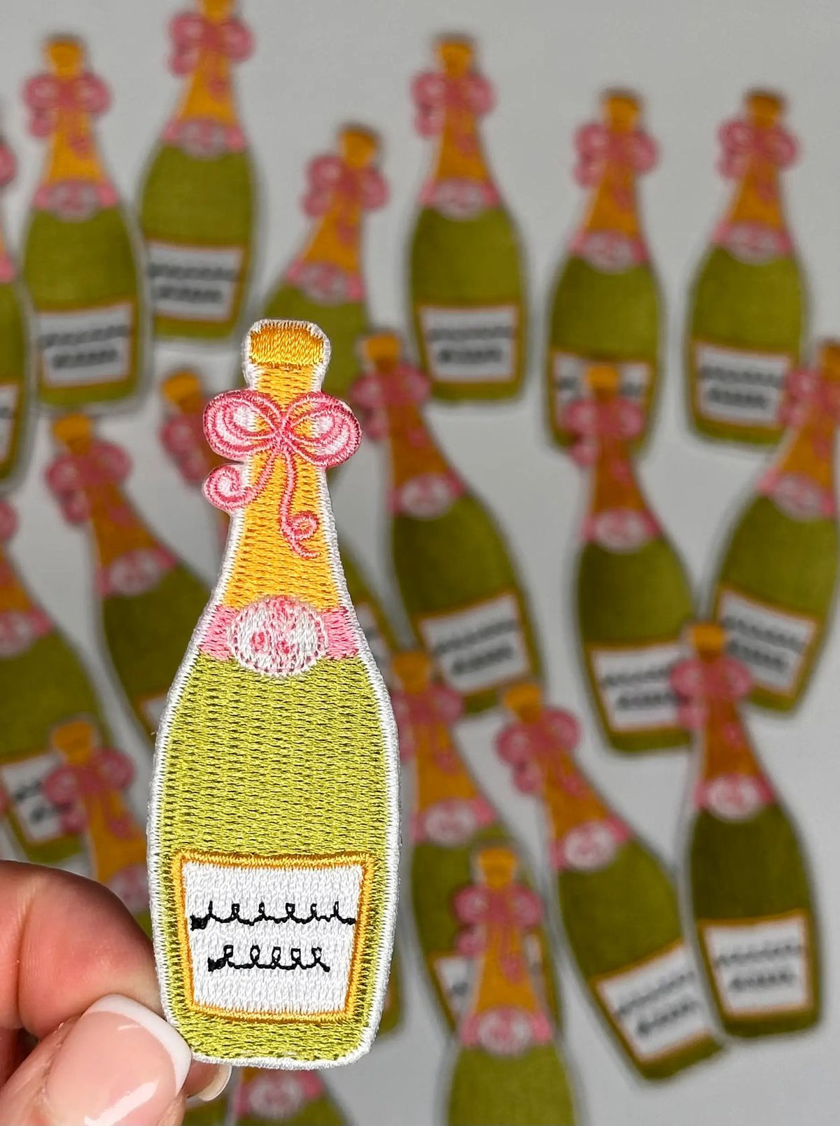 Patch of a champagne bottle with a pink bow held over multiple similar patches on a white background.