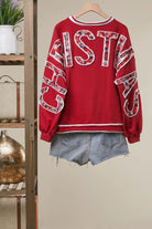 Back view of a red crewneck sweatshirt with CHRISTMAS spelled in plaid and floral patchwork letters across the back and sleeves, styled with denim shorts