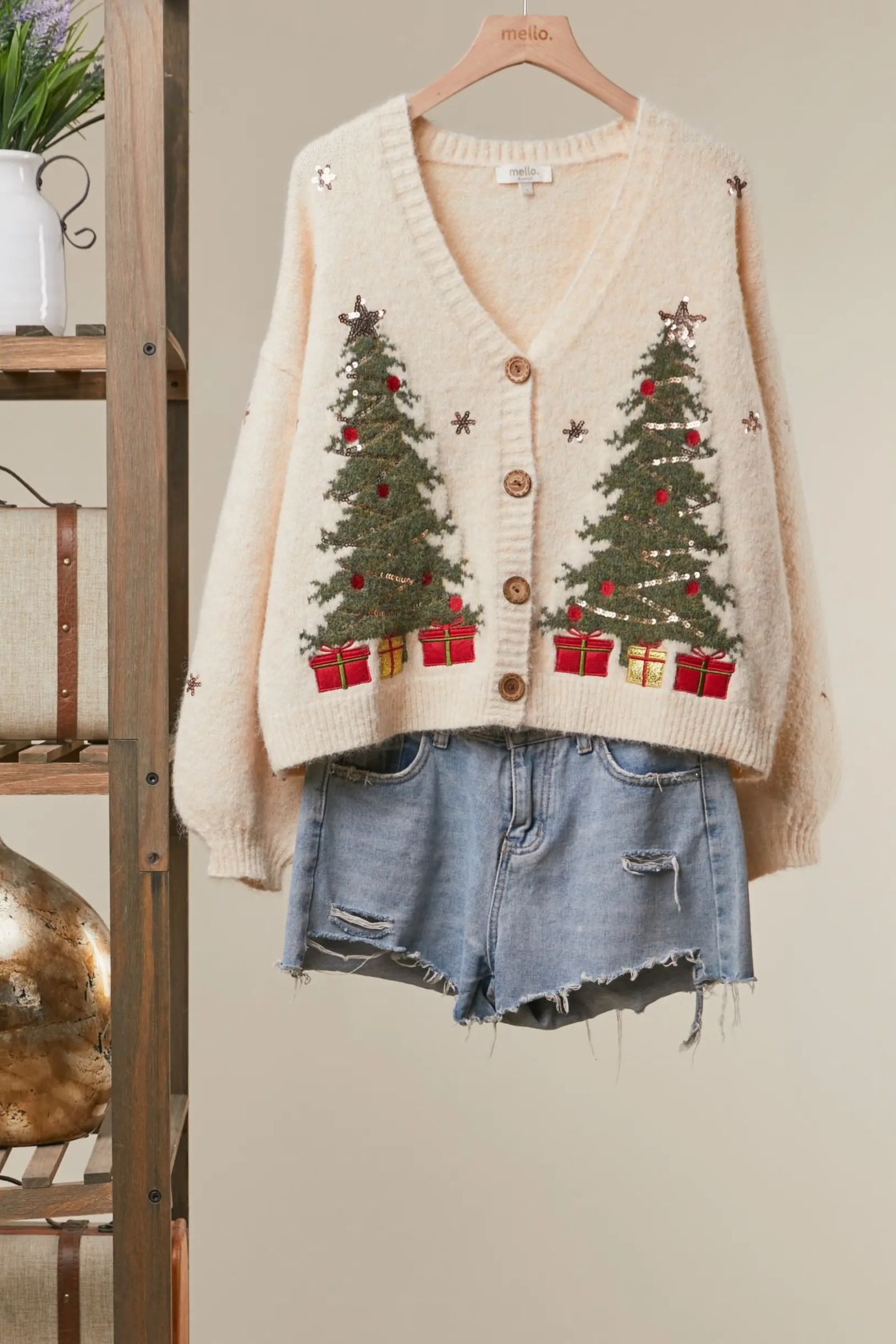 Ivory V-neck cardigan with embroidered green Christmas trees, red and gold gift boxes, and sequin snowflakes, styled with denim shorts on a wooden hanger