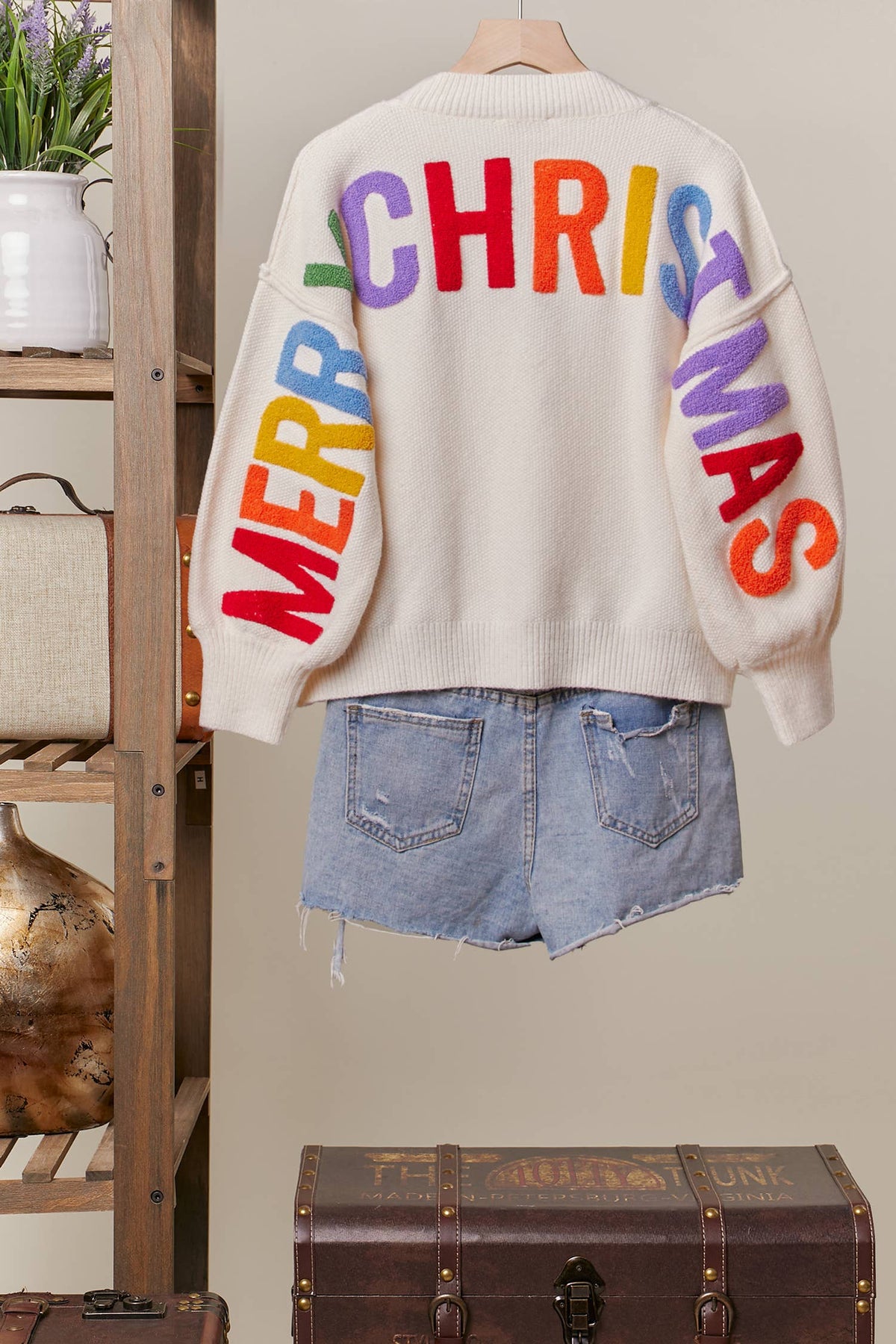 Back of the ivory Colorful Christmas Sweater with rainbow Merry Christmas lettering across the back and sleeves, on a hanger with denim shorts