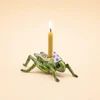 A novelty candle shaped like a green grasshopper with a lit yellow taper candle inserted, shown on a plain light beige background.