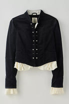A cropped black jacket with a high collar, decorative silver-tone snap buttons down the front, and a cream ruffled underlayer peeking out at the hem and cuffs, shown against a neutral light gray background.