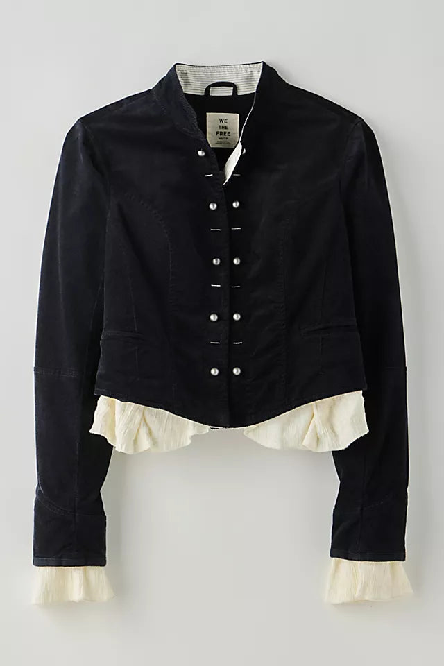A cropped black jacket with a high collar, decorative silver-tone snap buttons down the front, and a cream ruffled underlayer peeking out at the hem and cuffs, shown against a neutral light gray background.