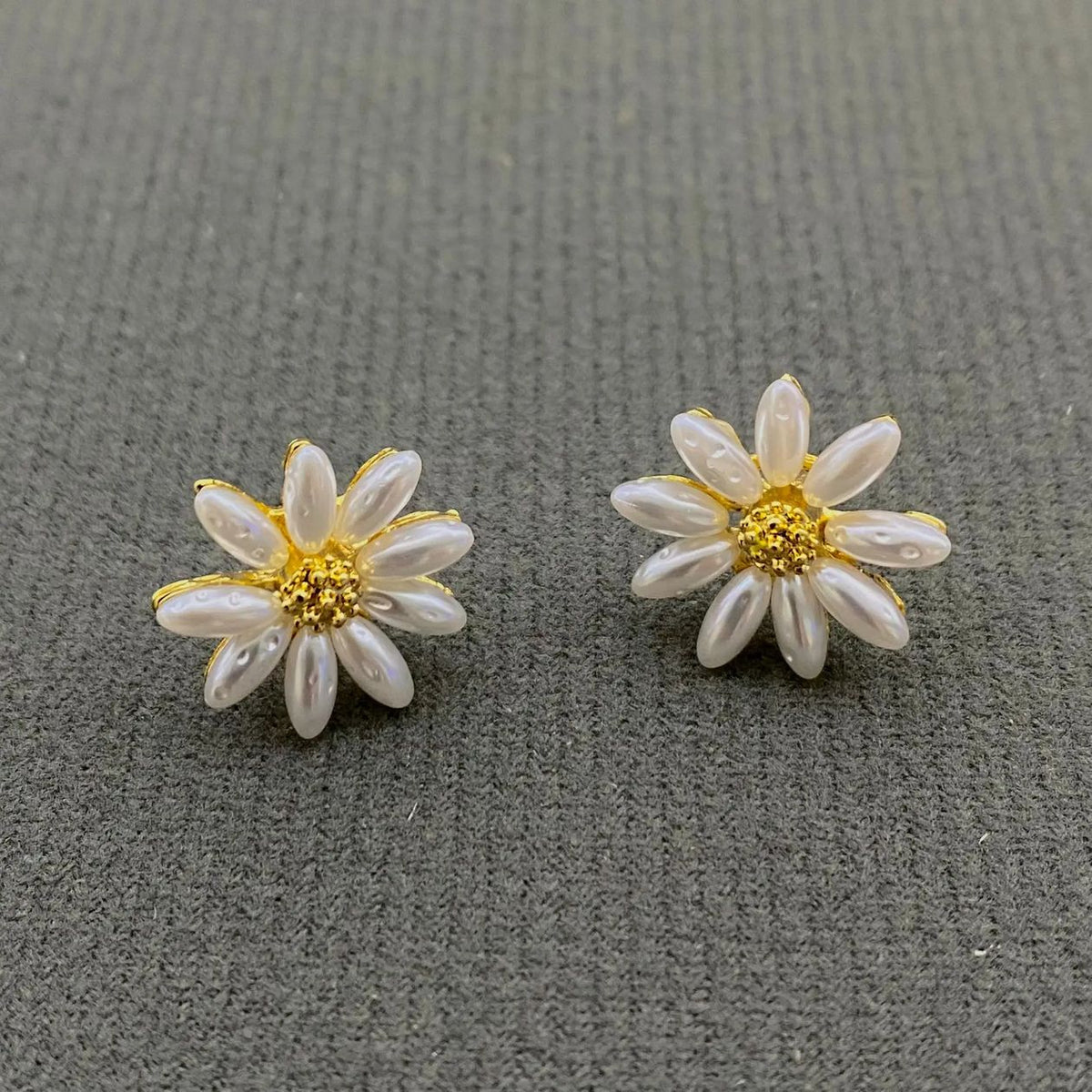 A pair of daisy flower stud earrings with white faux pearl petals and gold centers, displayed on a textured gray background.