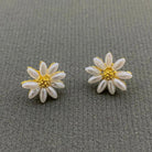 A pair of daisy flower stud earrings with white faux pearl petals and gold centers, displayed on a textured gray background.