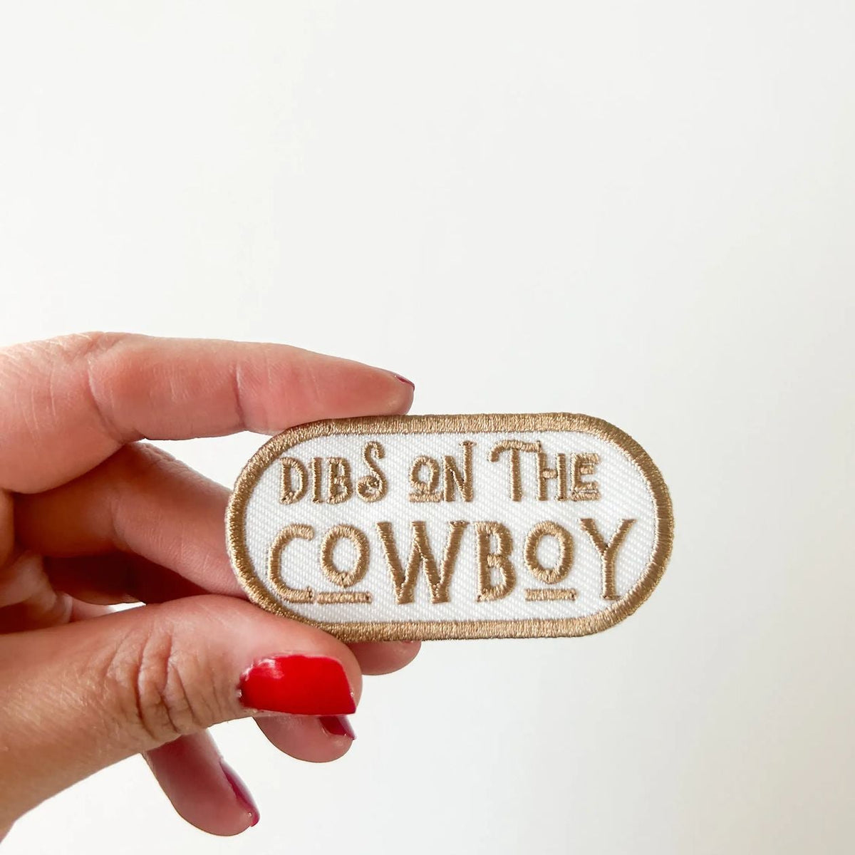 Hand holding a patch with 'Dibs on the Cowboy' text against a white background