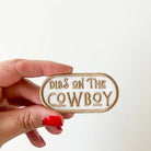 Hand holding a patch with 'Dibs on the Cowboy' text against a white background