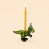 A green dinosaur-shaped candle holder with a lit yellow candle inserted into its back, set against a solid light beige background.