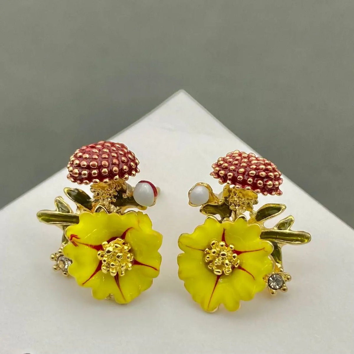 A pair of mushroom-shaped stud earrings with yellow flower petals, gold-toned stems and centers, and reddish-brown textured caps, displayed on a white triangular stand against a gray background.
