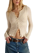 Model wearing the Free People Love Cardi, a fitted winter white diamond pointelle knit cardigan with ruffle trim, with low-rise jeans and a brown leather belt