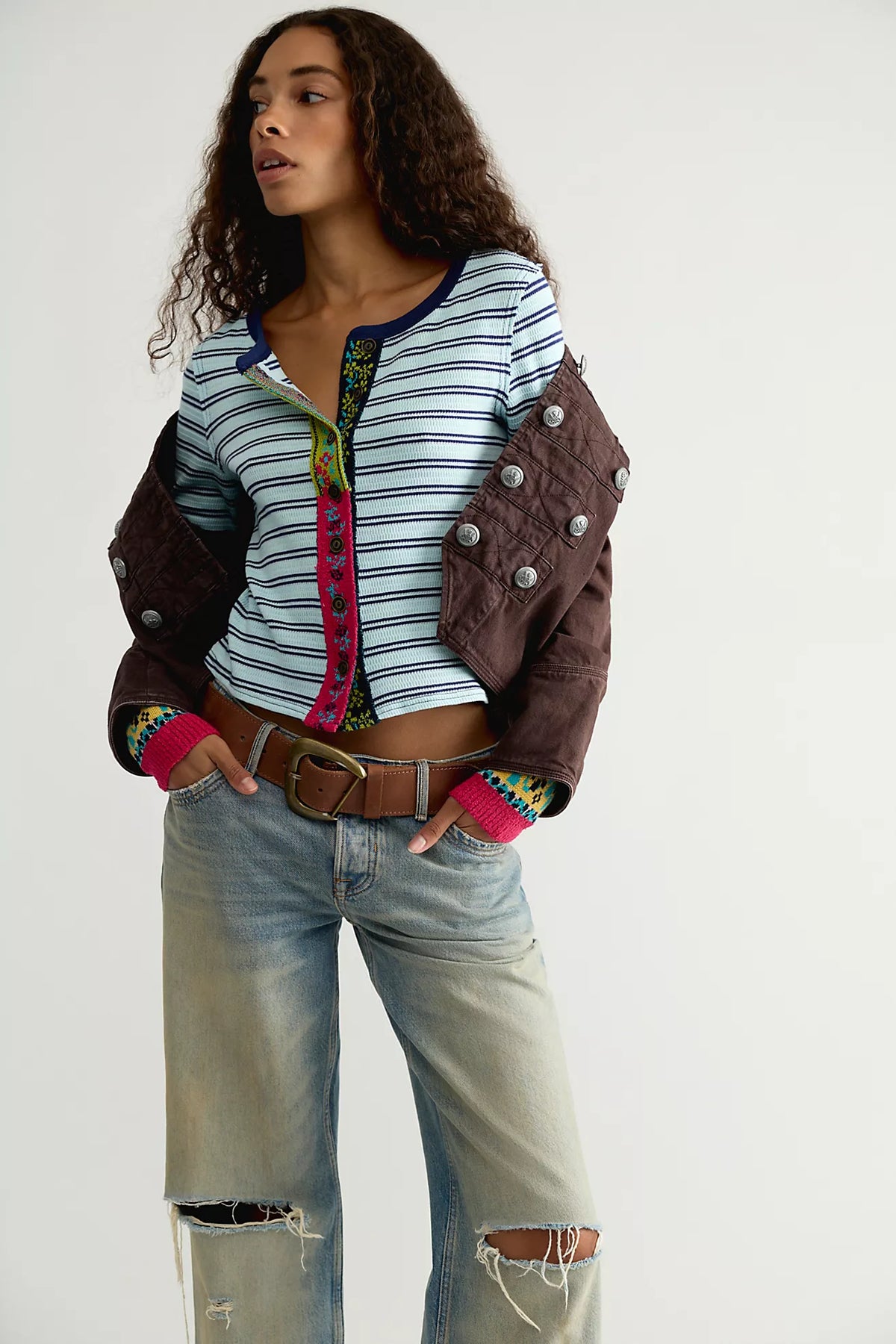 Model wearing the Free People Noa Cardi buttoned, a blue and navy striped cardigan with pink embroidered placket and cuffs, styled with a brown jacket over the shoulders and distressed jeans