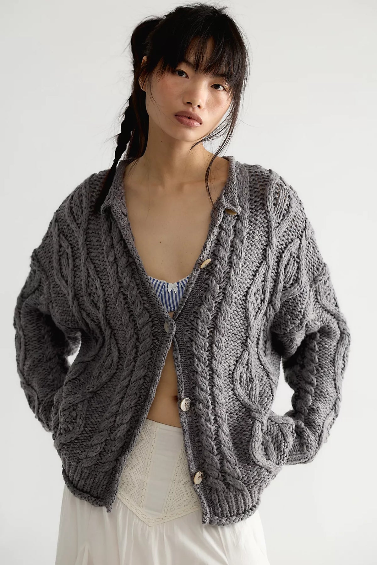 Model wearing the Free People Opal Cardigan, an oversized steel grey chunky cable knit cardigan with button front, over a striped top and white skirt