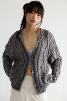 Model wearing the Free People Opal Cardigan, an oversized steel grey chunky cable knit cardigan with button front, over a striped top and white skirt