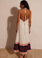 A woman shown from the back wearing a white halter-neck maxi dress with a tie-back neckline and a multicolor floral patterned hem, standing against a textured beige wall.