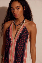 A woman wearing a sleeveless halter-style dress with a dark navy base, red and beige geometric border patterns, and a red floral print, styled with layered necklaces against a plain beige wall.