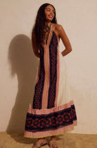 A woman modeling a sleeveless maxi dress with a bohemian patterned design in navy, pink, and cream, standing against a textured beige wall.