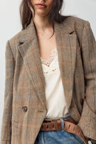 Model wearing the Free People Shiloh Blazer, a boxy brown and beige plaid wool-blend blazer, over a cream lace-trim cami with jeans and a brown leather belt