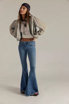 A model wearing a light gray oversized bomber jacket, a beige crop top, blue flare jeans, and a dark purple headband, standing against a plain light gray studio background.