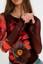 A close-up of a woman wearing a sheer long-sleeve top in dark brown with a large red and orange floral print and ruffled cuffs.