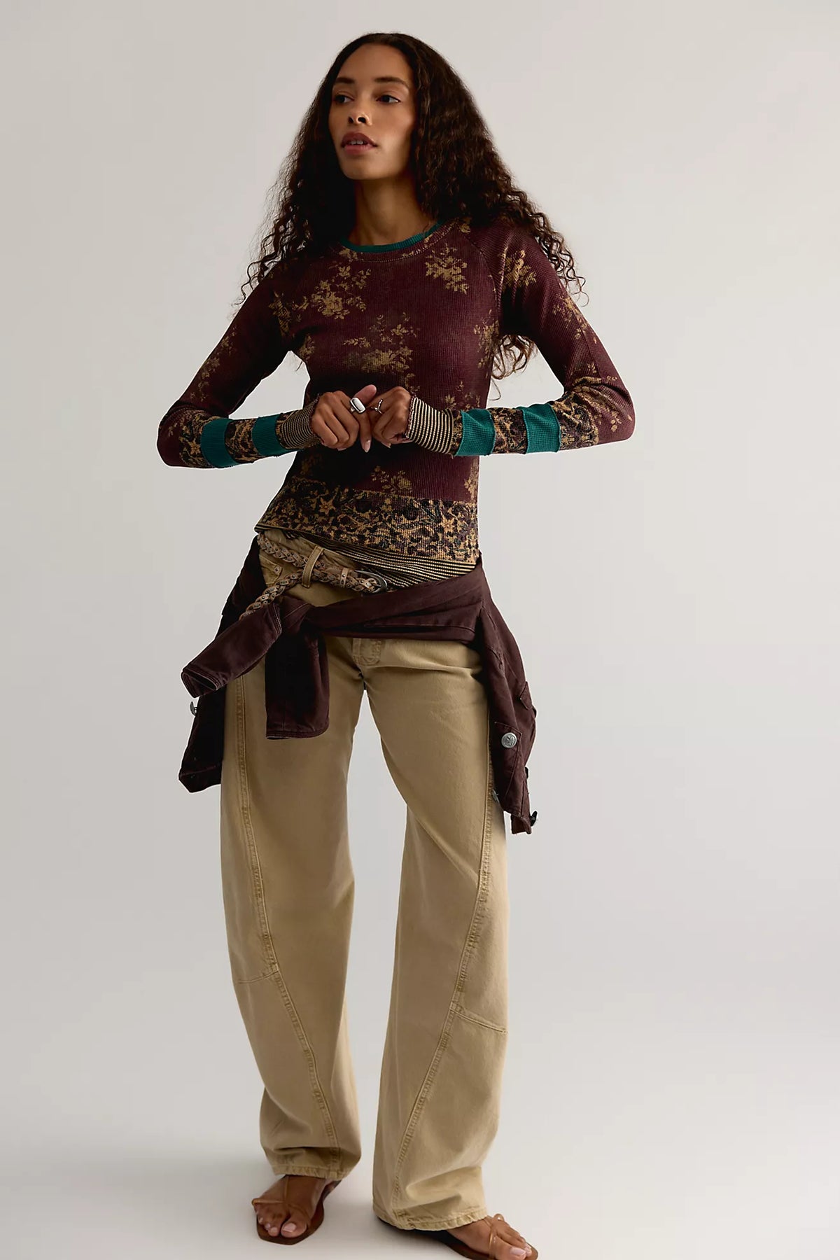 A woman modeling a long-sleeve maroon top with a gold floral pattern and teal cuffs, paired with beige pants and a dark brown jacket tied around her waist, against a light gray studio background.