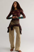 A woman modeling a long-sleeve maroon top with a gold floral pattern and teal cuffs, paired with beige pants and a dark brown jacket tied around her waist, against a light gray studio background.