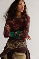 A woman modeling a long-sleeve crewneck top in deep burgundy with a gold floral pattern, teal ribbed cuffs, and a matching patterned waist sash, paired with khaki pants against a light gray studio background.