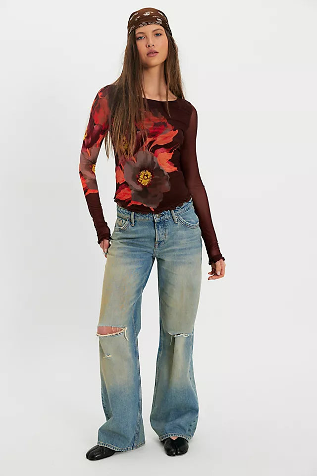 A woman modeling a long-sleeve top with a dark red base and a large red and orange floral print on the front, paired with light-wash distressed jeans and a brown cap, against a plain white studio background.