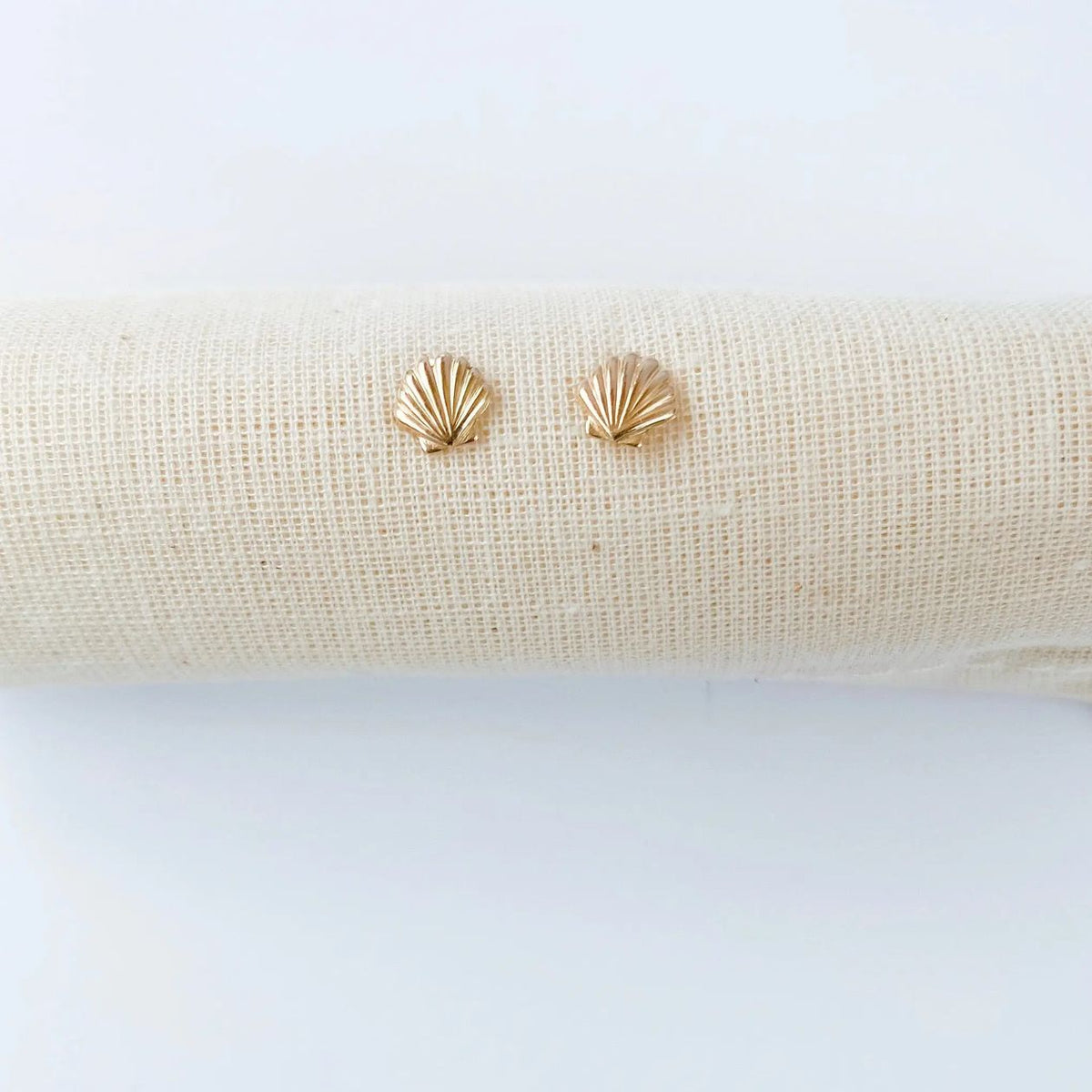 A pair of small gold stud earrings shaped like scallop shells, displayed on a textured beige fabric roll against a clean white background.