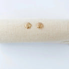 A pair of small gold stud earrings shaped like scallop shells, displayed on a textured beige fabric roll against a clean white background.