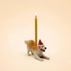 A small figurine of a brown and white dog wearing a red Santa hat, standing next to a single lit yellow candle on a plain beige background.