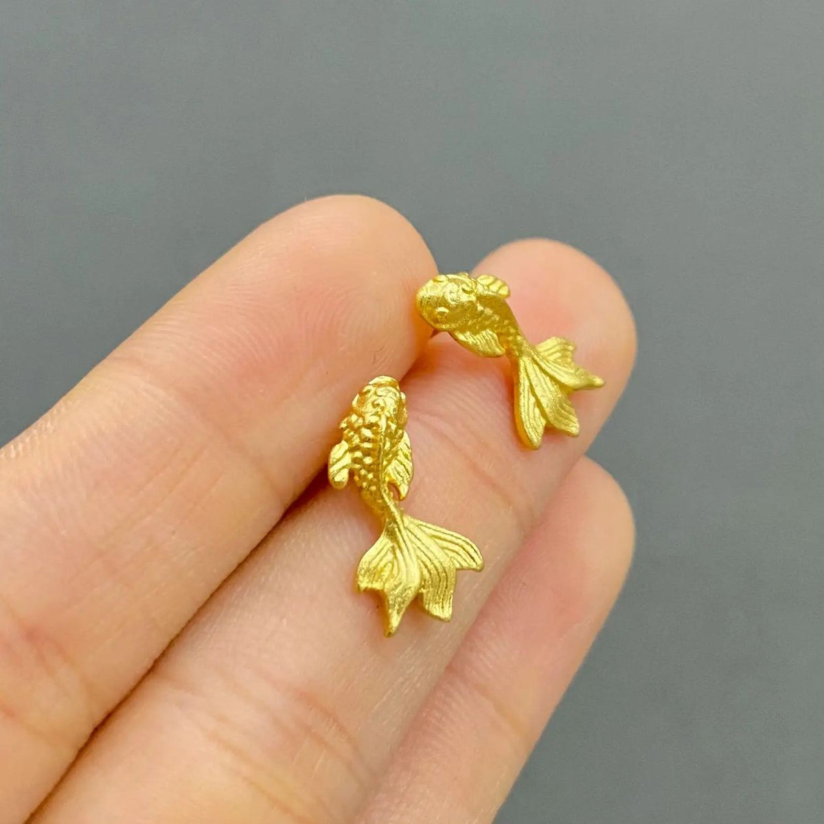 A pair of small gold fish-shaped stud earrings held between two fingers against a plain gray background.