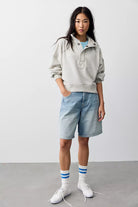 A model stands in a studio wearing a light gray oversized hoodie, light-wash denim shorts, white crew socks with blue stripes, and white sneakers against a plain light gray background.