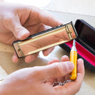 Person cleaning a gold harmonica with a yellow brush on a light wooden surface.