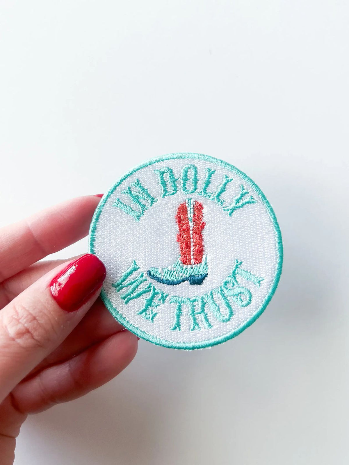 Circular embroidered patch with 'In dolly We Trust' text held by a hand on a light background