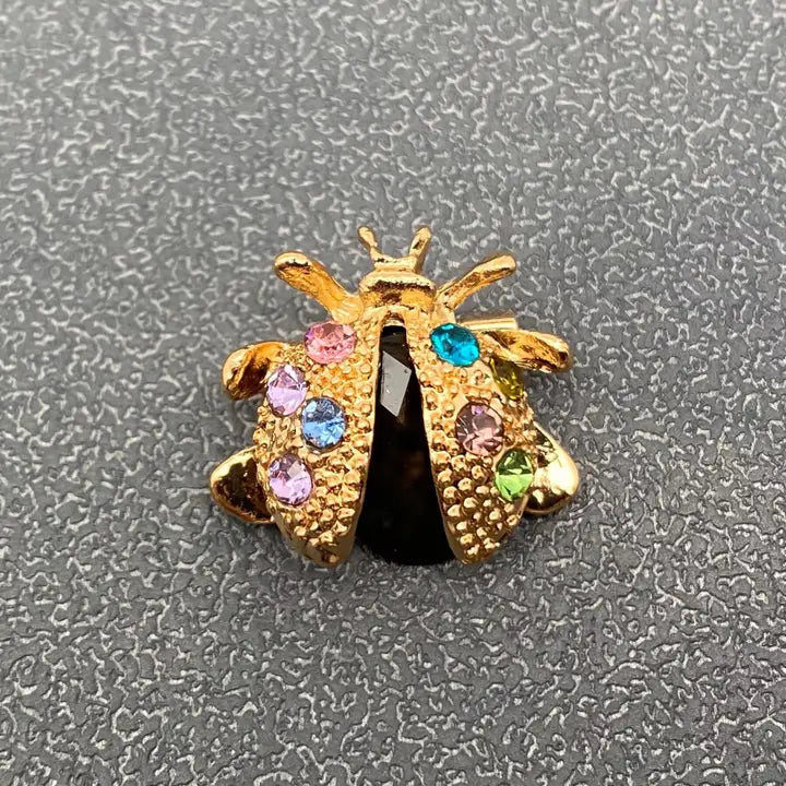 A gold ladybug-shaped brooch or charm embellished with multiple colorful rhinestones in pink, blue, green, and purple, set against a textured gray background.