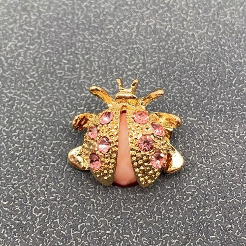 A gold ladybug-shaped brooch embellished with pink rhinestones and a pink center stripe, photographed on a textured gray background.